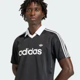 Camiseta Adidas Originals de manga corta, color negro con sutiles rayas verticales, cuello polo en V de color blanco, al igual que las líneas características de la marca en los hombros. Cuenta con el logotipo de la marca y la inscripción "adidas" en el pecho en color blanco.