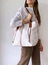 Cartera tipo tote de cuero vacuno con acabado texturizado en tono blanco hueso. Presenta detalles de costuras artesanales con tientos de cuero marrón en los bordes y correas de hombro a juego.
