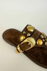 Sandalia plana de gamuza marrón, estilo ojota, con apliques metálicos dorados y hebilla decorativa.