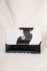 Billetera pequeña con solapa de cuero vacuno con pelo en estampado animal print blanco y negro, y cuerpo inferior de cuero liso negro. Incluye un botón metálico decorativo en el centro de la solapa.