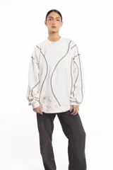 Buzo blanco de manga larga con diseño holgado, cuello redondo y costuras externas visibles en contraste. Cuenta con una etiqueta decorativa en el frente inferior.