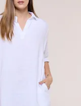 Vestido corto blanco de algodón, marca Dash, con cuello camisero, escote en V y bolsillos laterales. Presenta mangas tres cuartos y calce holgado con ruedo irregular.