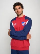 Buzo de felpa con diseño de bloques de color, predominantemente rojo en el cuerpo y azul marino en las mangas y hombros. Presenta el escudo del Club Nacional de Football y el logo de Umbro en el pecho.