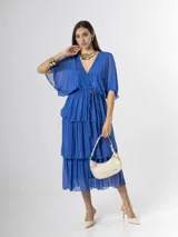 Vestido midi de seda italiana color azul, con escote en V, mangas murciélago amplias, falda de tres volados y cintura elastizada con lazo ajustable al tono y hebilla circular dorada.