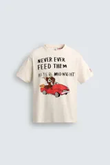 Camiseta de corte relajado color crudo, con cuello redondo y manga corta. Presenta un estampado frontal con una ilustración de Gizmo de la película Gremlins conduciendo un auto rojo y el texto 'Never ever feed them after midnight'.