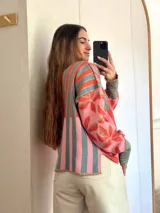 Sweater de tejido de punto con silueta oversize, cuello redondo y mangas largas abullonadas. Presenta un diseño patchwork con rayas verticales en tonos rojo, rosa y gris verdoso en el cuerpo, y un estampado floral en tonos rosa y rojo con detalles dorados en las mangas. Los puños y el cuello tienen un ribete a rayas horizontales.