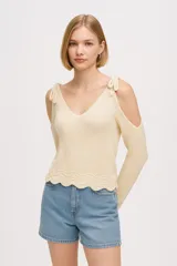 Sweater de tejido de punto color natural, con diseño de hombros descubiertos, mangas largas y detalle de lazos anudados en los hombros. Presenta un cuello en V profundo y un borde inferior con terminación ondulada.