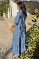 Vestido largo de estilo camisero en color azul, con cuello clásico, cierre frontal de botones y mangas tres cuartos con trabillas. Presenta un diseño de falda con cortes horizontales que le otorgan volumen y movimiento.
