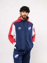 Campera deportiva con cierre frontal y capucha, diseñada con bloques de color en azul marino, rojo y blanco. Presenta el escudo del Club Nacional de Football y el logo de Umbro en el pecho.