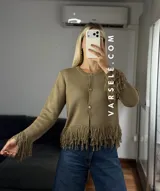 Cárdigan de tejido de punto color crema, con cuello redondo, mangas largas y cierre frontal con botones metálicos. Presenta detalles de flecos en los puños y en el ruedo.
