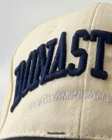 Gorra de béisbol beige con visera y botón superior azul marino. Presenta bordado frontal en azul marino con la inscripción "Bonastun South American League".