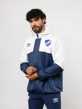 Campera de lluvia Umbro del Club Nacional de Football, con diseño de bloques de color en rojo y azul marino. Presenta capucha, cierre frontal, puños elásticos y detalles de piping blanco. Incluye logo de Umbro y escudo del club en el pecho.