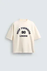 Sudadera boxy fit de color beige con interior de rizo, cuello redondo y manga corta. Presenta un estampado contrastante en el frente con la inscripción "Deep Founders London 90" y acabados irregulares en los bordes.