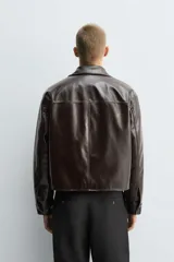 Cazadora cropped de hombre confeccionada en tejido efecto piel de acabado brillante y color marrón. Cuenta con cuello de solapas de muesca, manga larga con puños abotonados, bolsillos de vivo en la cadera y cierre frontal con botonadura.