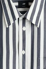 Camisa de popelina de algodón con diseño de rayas verticales en azul y blanco, cuello inglés, cierre frontal de botones y un bolsillo en el pecho. Presenta mangas largas con puños abotonados y corte relaxed fit.