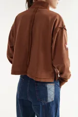 Buzo rústico marrón con cuello polera, mangas largas y calce boxy. Presenta costuras externas decorativas, puños acanalados y un bordado sutil en el pecho.