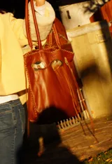 Cartera de cuero color camel, de silueta amplia y blanda, con manijas largas y terminación artesanal en los bordes. Cuenta con apliques decorativos frontales de alpaca y nácar, tientos largos colgantes y bolsillo interno.