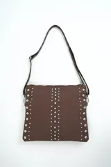 Cartera bandolera marrón decorada con tachas metálicas plateadas en los laterales y en dos líneas verticales frontales, con correa ajustable y cierre superior.