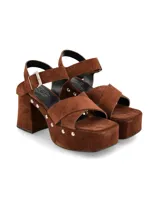 Sandalia de plataforma de gamuza color marrón, con taco ancho, tiras cruzadas en la parte delantera y tira ajustable al tobillo con hebilla metálica. Cuenta con detalles de tachas plateadas en el lateral de la suela.