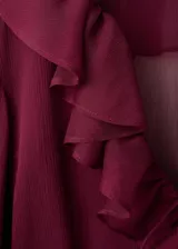 Blusa bordó de tejido fluido y semi transparente, con escote en V pronunciado, mangas largas abullonadas con volantes en los puños y detalles de volantes en el cuello y el ruedo.