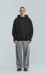 Hoodie negro de frisa premium, corte oversize, con capucha ajustable y puños y cintura elastizados. Presenta un bolsillo canguro frontal y un detalle de etiqueta tejida con texto repetido en los cordones de la capucha y en la manga izquierda.