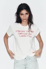 Camiseta blanca de algodón flamé, con cuello redondo, manga corta y estampado frontal de texto en color rojo con la frase 'Starting the day confused but committed'.
