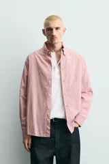 Camisa de corte relajado confeccionada en tejido de algodón, con diseño de rayas verticales en tonos rosados y blancos. Presenta cuello abotonado, cierre frontal de botones, manga larga con puños abotonados y bajo asimétrico.