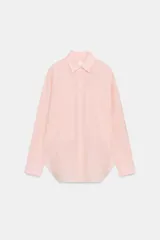 Camisa de corte oversize color rosa claro con diseño de rayas verticales sutiles. Presenta cuello solapa desmontable, cierre frontal con botones ocultos bajo solapa, mangas largas con puños y bajo asimétrico redondeado. Incluye dos bolsillos frontales amplios.