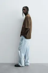 Camiseta de algodón color marrón oscuro, corte relajado (relaxed fit) y manga corta. Presenta un estampado frontal con un lobo o cánido corriendo, el texto "The Riders Project", "VISION & TEMPER" y "0.79 per gallon" en tonos grises y rosados.