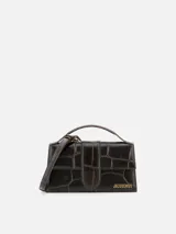 Bolso Le Grand Bambino de Jacquemus confeccionado en cuero de ternera con estampado efecto cocodrilo en color marrón oscuro. Presenta una silueta estructurada, solapa frontal con cierre magnético y tira vertical, asa superior rígida y correa ajustable y desmontable. Incluye detalles metálicos dorados con el logotipo de la marca en el frente.