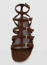 Sandalias planas de gamuza marrón con diseño de múltiples tiras cruzadas y hebillas metálicas doradas. Cuenta con cierre de hebilla en el tobillo.