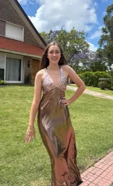 Vestido largo con estampado tie-dye en tonos violeta y fucsia, escote en V con detalles bordados dorados, cut-outs laterales bajo el busto y tirantes finos que se cruzan y atan en la espalda.