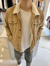 Chaqueta de pana color marrón tostado, con forro interno y cuello de corderito sintético blanco. Presenta cierre frontal de botones metálicos, bolsillos frontales con solapa en el pecho y bolsillos laterales.