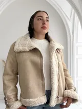 Chaqueta color beige con interior de piel sintética blanca, cierre cruzado con cremallera y detalles de hebillas en los puños.