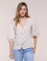Blusa color beige confeccionada en lino, con escote en V pronunciado y cierre frontal de múltiples lazos para atar. Presenta mangas abullonadas tres cuartos con puños elásticos y corte relajado.