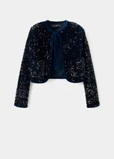 Chaqueta crop de terciopelo con lentejuelas, cuello redondo, manga larga y cierre de lazo. Cuenta con dos bolsillos de parche frontales y forro interior.