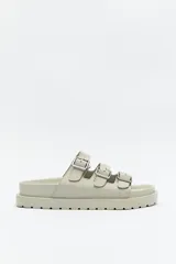 Sandalia tipo slide de color beige claro, con tres tiras ajustables sobre el empeine, cada una con hebilla metálica plateada. Posee una plataforma robusta con relieve en la suela.