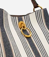 Cartera bucket pequeña de Satinato con diseño de rayas verticales en tonos blanco, azul marino y beige, confeccionada en tejido estructurado. Cuenta con asa de mano fija, asa larga desmontable de cuero sintético marrón, detalles de herrajes metálicos dorados, argolla metálica colgante en el frente y cierre superior.