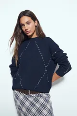 Sweater de tejido suave color marrón con diseño de líneas bordadas en color rojo. Presenta cuello redondo, manga larga y calce relajado.