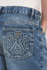 Jeans de denim azul con calce holgado y tiro medio, con diseño de cintura asimétrica cruzada, roturas localizadas y ruedo desflecado con degradé.