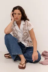 Camisa blanca de manga corta con cuello solapa, cierre frontal de botones y diseño bordado en hilo azul marino con motivos florales y vegetales.