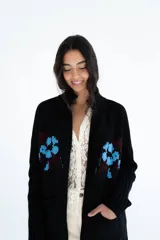 Cardigan de punto color marrón con cierre frontal, cuello alto y bolsillos aplicados. Presenta diseño tejido localizado con detalles en violeta y negro en el pecho.