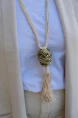 Collar largo con cordones de algodón color crema, que presenta un dije central esférico formado por hilos metálicos entrelazados en tonos dorados y plateados, finalizando con una borla de flecos de algodón.