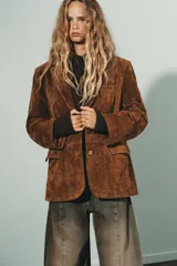 Blazer de gamuza color marrón con cuello solapa, manga larga con hombreras y cierre frontal con botones. Incluye bolsillos con solapa en el delantero, bolsillo de vivo en el pecho y cinturón de lazada en el mismo material.