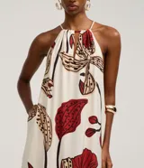 Vestido midi de corte evasé, con escote halter y abertura frontal en el busto, anudado en la parte superior. Presenta un estampado grande de flores y hojas en tonos rojo oscuro, beige y negro sobre fondo crudo. El diseño incluye un volante fruncido en el ruedo inferior.