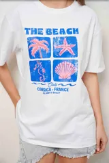Remeron de algodón blanco con cuello redondo y mangas cortas. Presenta un estampado frontal con la inscripción 'THE BEACH CLUB CORSICA FRANCE' y gráficos playeros en tonos azules y rosados.