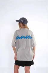 Remera oversize de algodón color gris claro, con cuello redondo y manga corta. Presenta un pequeño logo bordado en azul en el pecho.