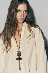 Collar largo con cadena de eslabones dorados y cuentas redondas en tonos rojo oscuro y verde azulado. El dije principal es una pieza metálica dorada con relieve de un pájaro, adornado con pequeñas piedras de colores, y un colgante de borla de cadena metálica oscura, unido a una pieza rectangular de resina color borgoña.