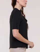 Blusa blanca de corte recto y tejido liviano de gasa con forro interno delantero. Presenta escote en V con cuello redondo y lazo central para anudar, mangas cortas con hilera de botones forrados en los laterales y ruedo ligeramente redondeado.