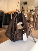 Cartera tipo hobo de textura gamuzada en color marrón oscuro, con diseño flexible y asa larga regulable. Presenta un detalle de flecos largos desmontables en uno de los laterales y cierre superior.